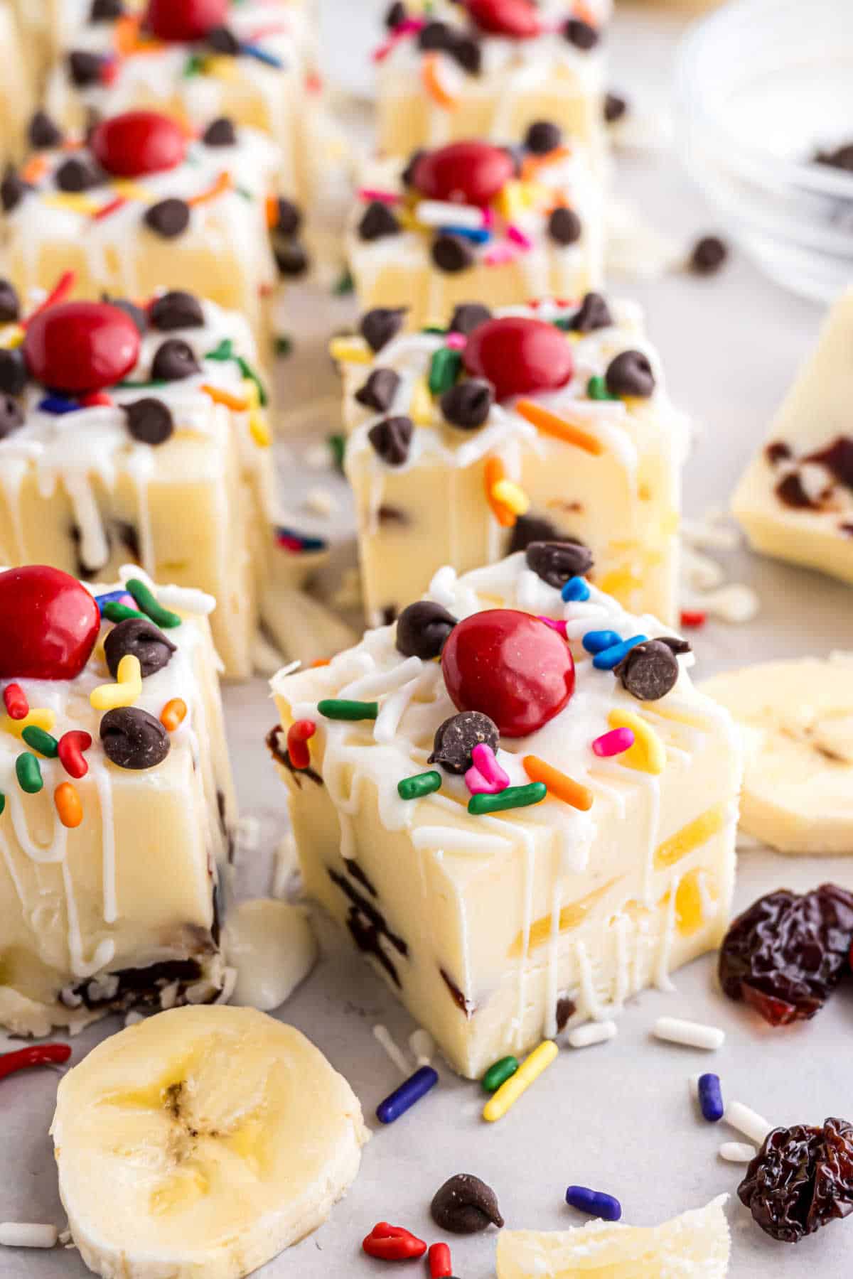 Indulge in Banana Split Fudge—white fudge squares topped with whipped cream, sprinkles, cherries, and chocolate chips, complete with banana slices nearby.