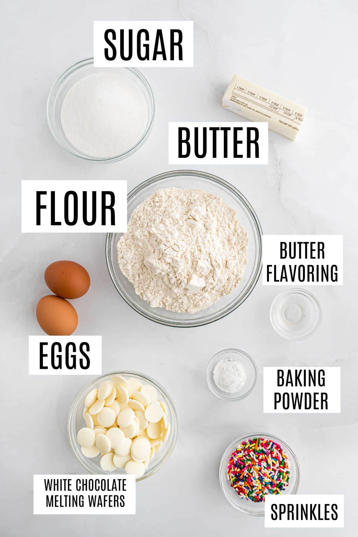 Top-down view of labeled ingredients for baking birthday cake biscotti: sugar, butter, flour, eggs, white chocolate, sprinkles, and baking powder.