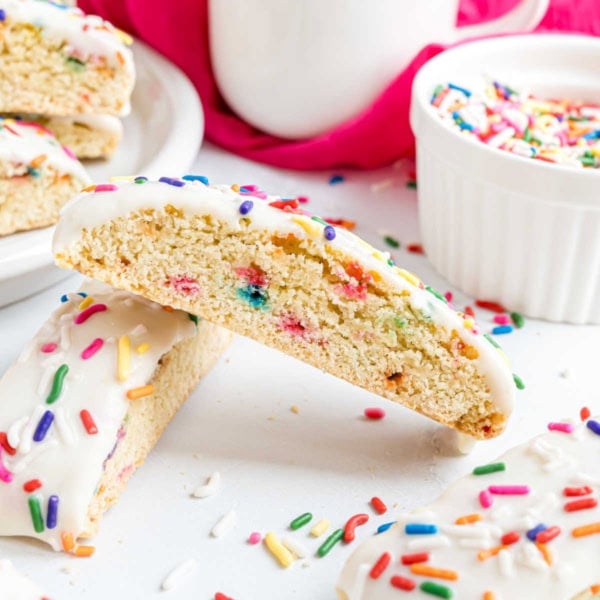 Birthday cake biscotti with white icing and colorful sprinkles, next to a bowl of sprinkles and a mug.
