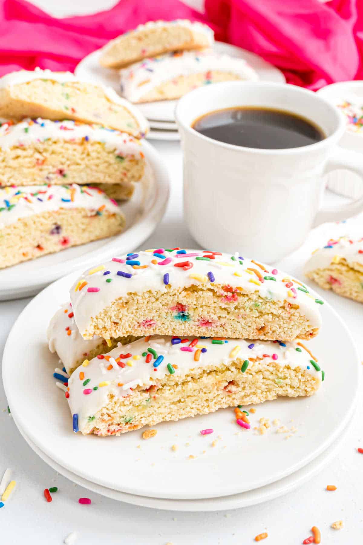 Two birthday cake biscotti with white icing and colorful sprinkles on a plate, next to a cup of black coffee.