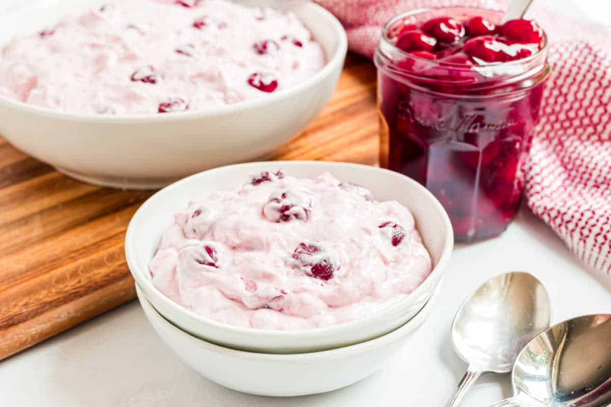 Bowl of creamy cherry dump salad dessert beside a jar of cherries, spoons, and a red patterned towel.