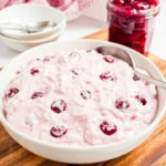 A bowl of pink cherry dump salad with a spoon sits beside a jar of cherries on a wooden board.