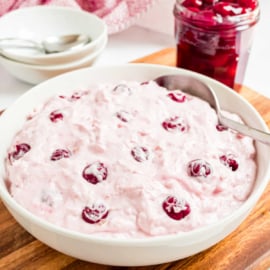 A bowl of pink cherry dump salad with a spoon sits beside a jar of cherries on a wooden board.