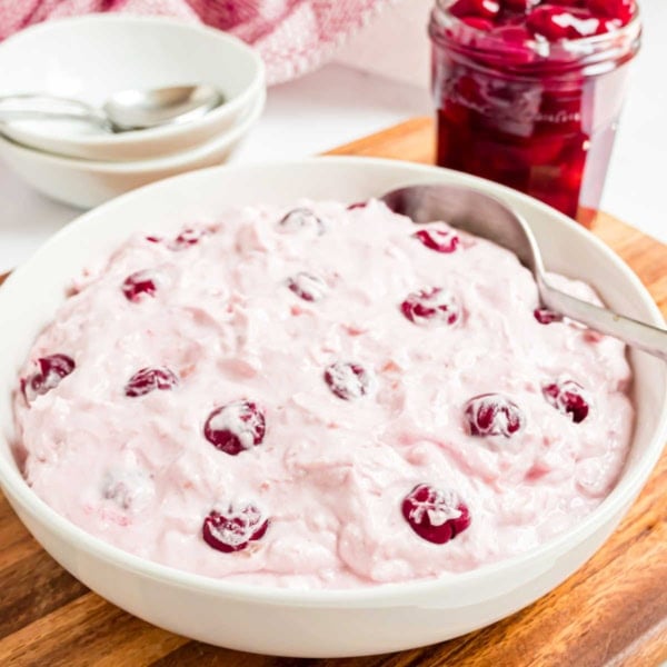 A bowl of pink cherry dump salad with a spoon sits beside a jar of cherries on a wooden board.