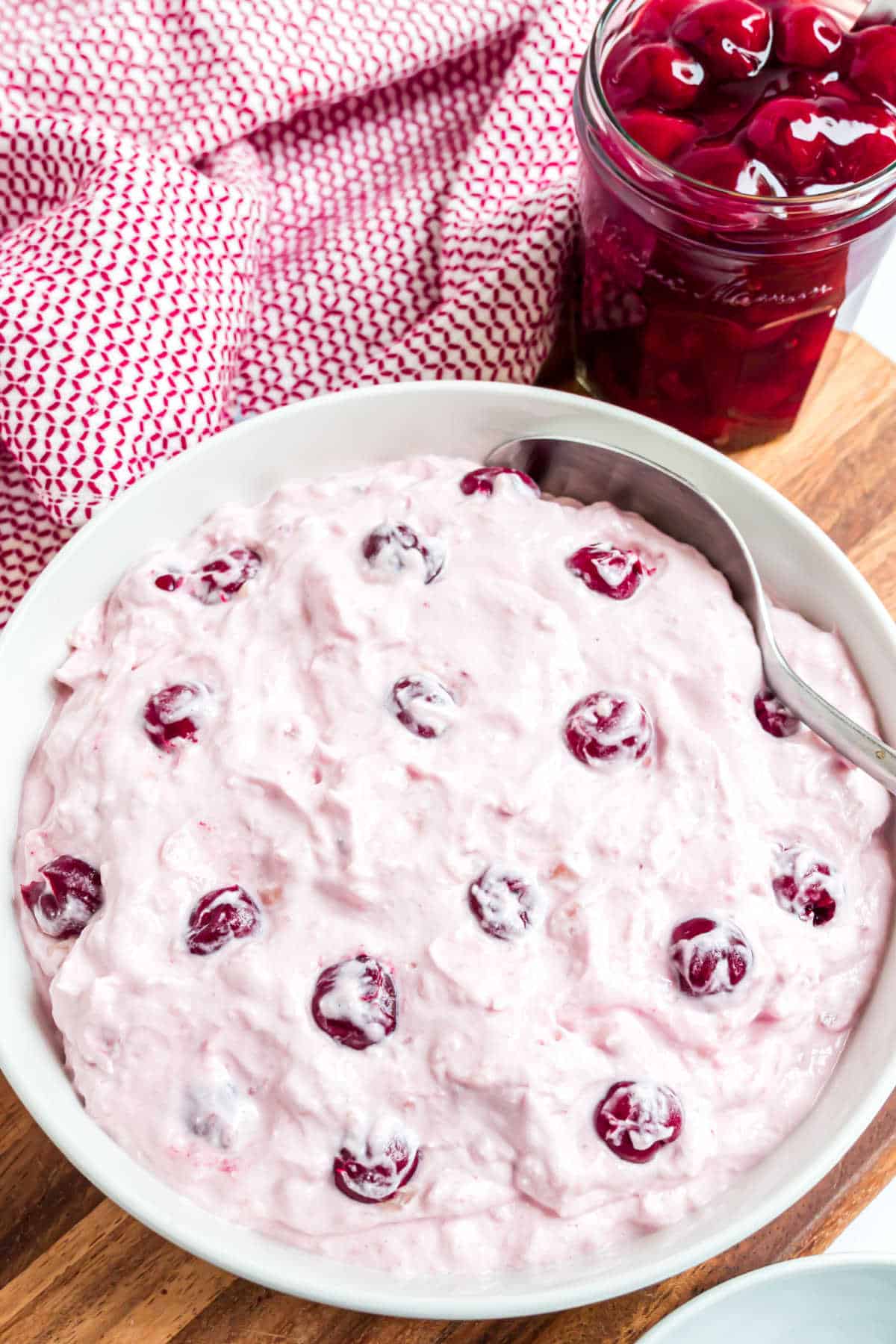 A bowl of creamy pink cherry dump salad with whole cherries sits beside a jar of cherries and a red patterned napkin.
