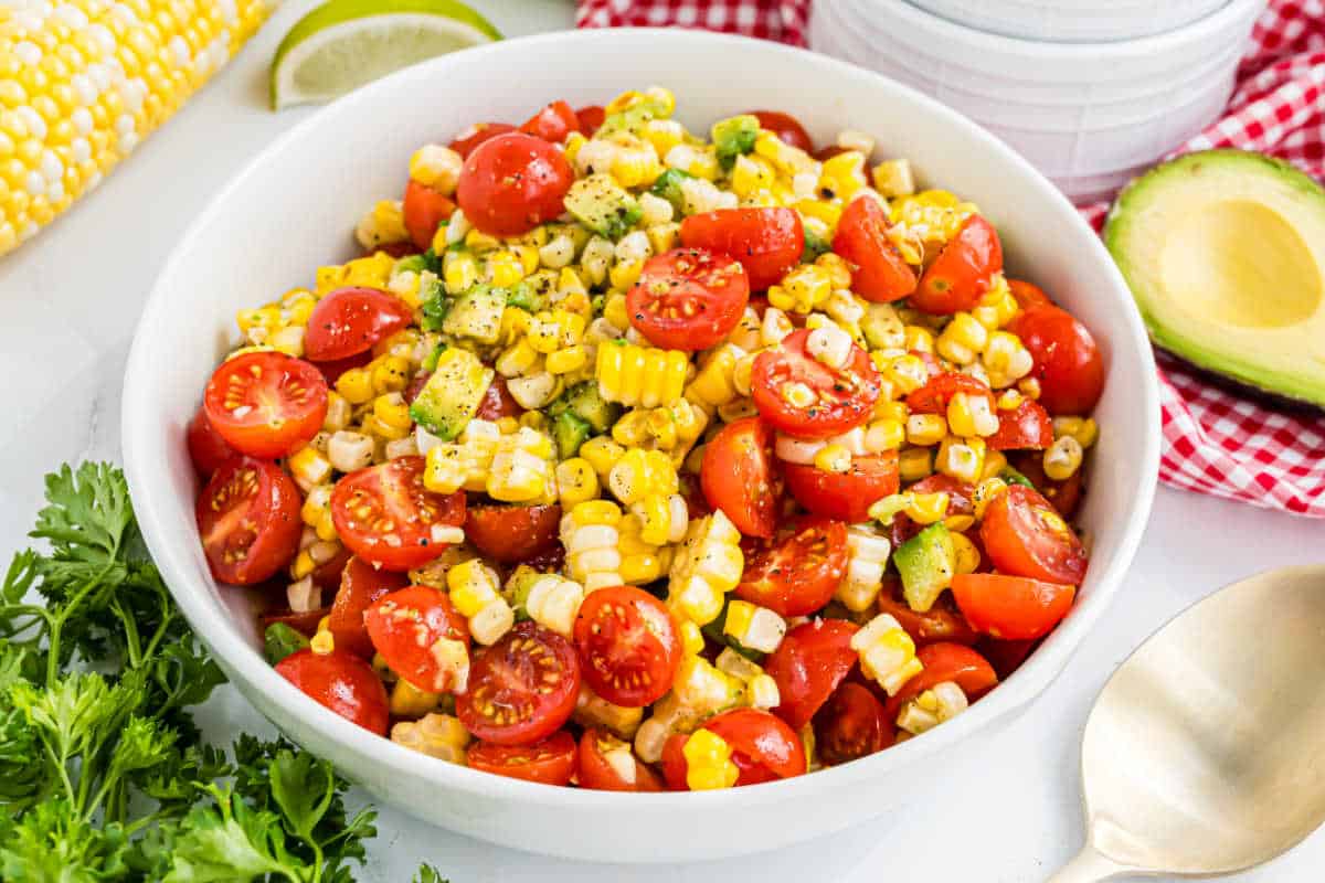 White bowl filled with roasted corn salad featuring cherry tomatoes and avocado, surrounded by fresh herbs and a sliced avocado.