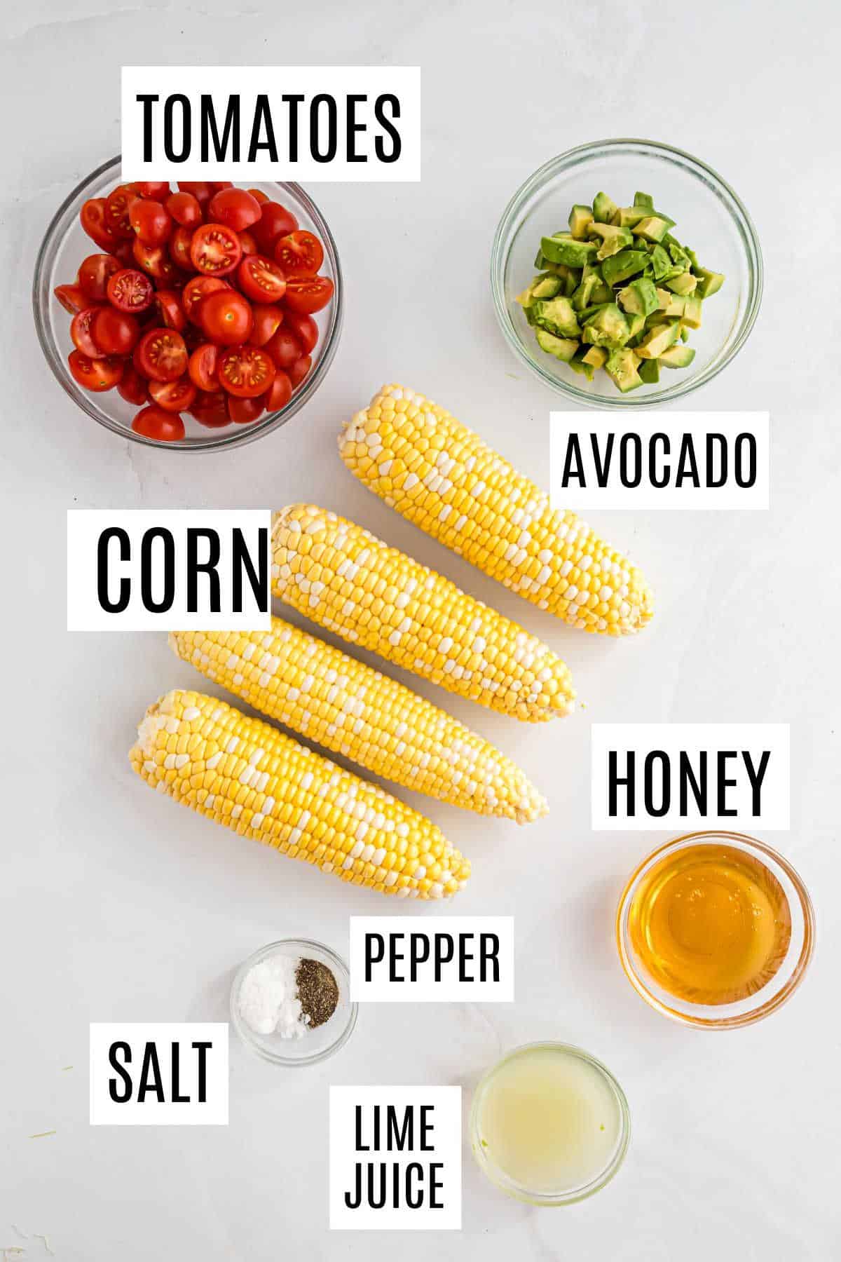 Ingredients for roasted corn salad: tomatoes, avocado, corn, honey, salt, pepper, and lime juice arranged on a white surface.