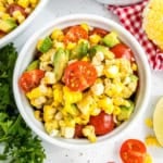 Bowl of roasted corn salad with cherry tomatoes, avocado, and fresh herbs on a white surface.