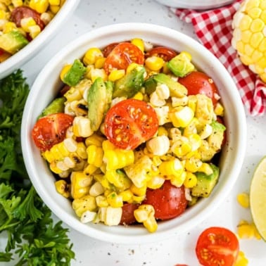 Bowl of roasted corn salad with cherry tomatoes, avocado, and fresh herbs on a white surface.