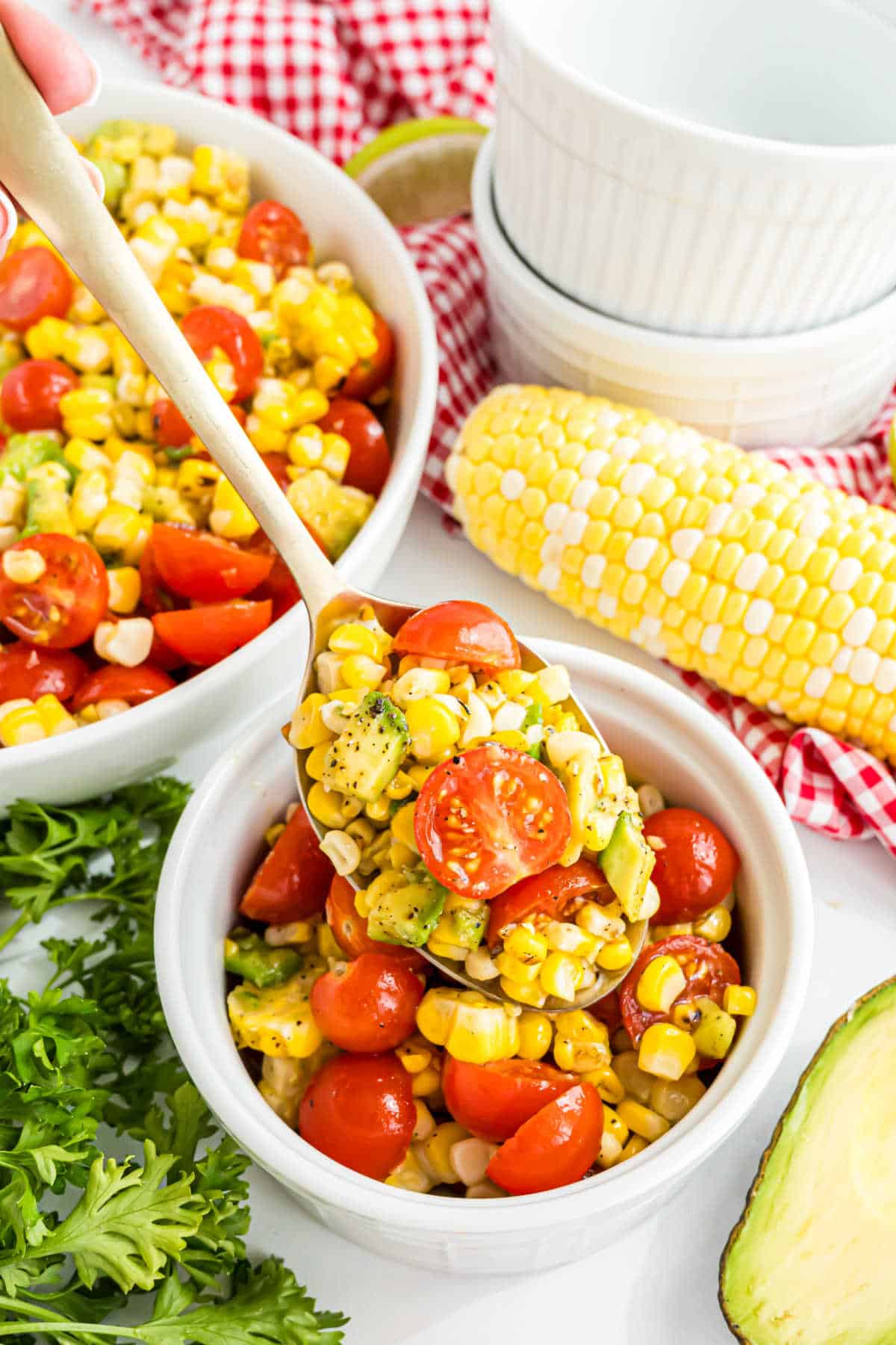A spoonful of roasted corn salad with cherry tomato and avocado over a bowl, with fresh corn and parsley nearby.