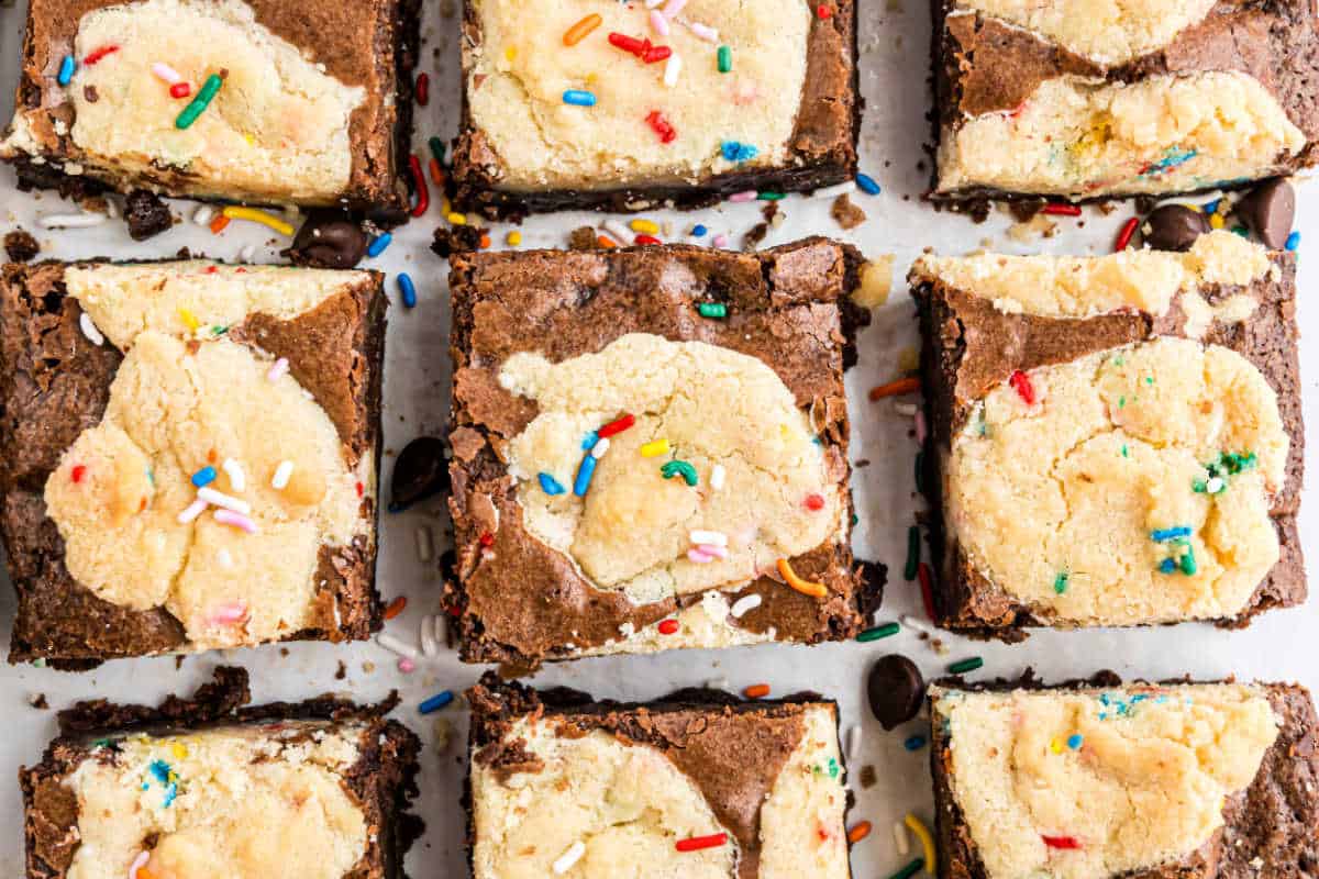 Nine sugar brookies topped with swirls of cheesecake and colorful sprinkles, arranged on a white surface.
