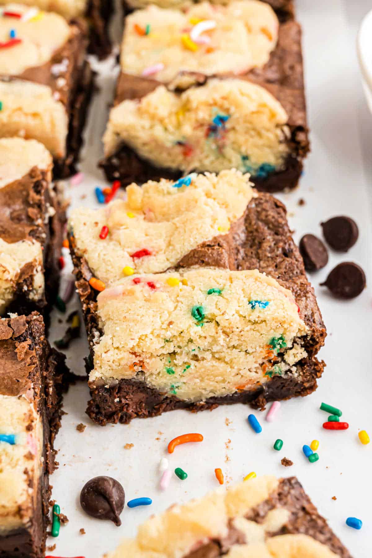 Sugar brookies topped with colorful sprinkles and chocolate chip cookie dough, cut into squares on a white surface.