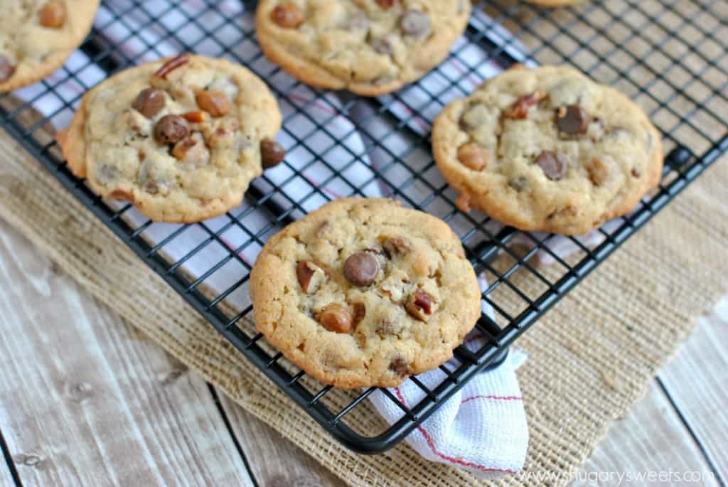 Chocolate chip cookies on a wire rack with chocolate chips, pecans, and caramel.