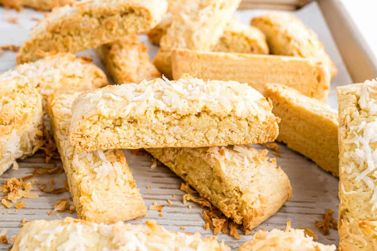 Coconut biscotti stacked on a baking sheet lined with parchment paper, each piece topped with shredded coconut.