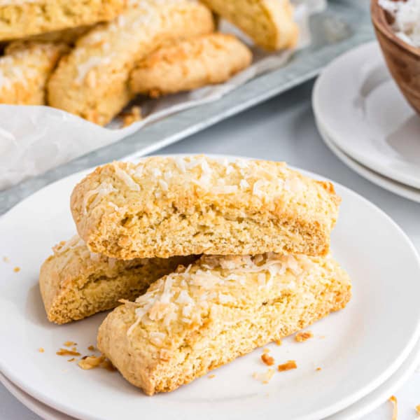 Two coconut biscotti stacked on a white plate, with extra coconut biscotti arranged in the background.