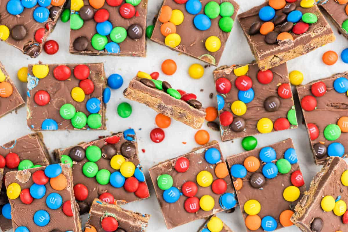 Squares of chocolate candy bark topped with colorful M&M candies resemble rainbow fudge cookie bars scattered on a white surface.