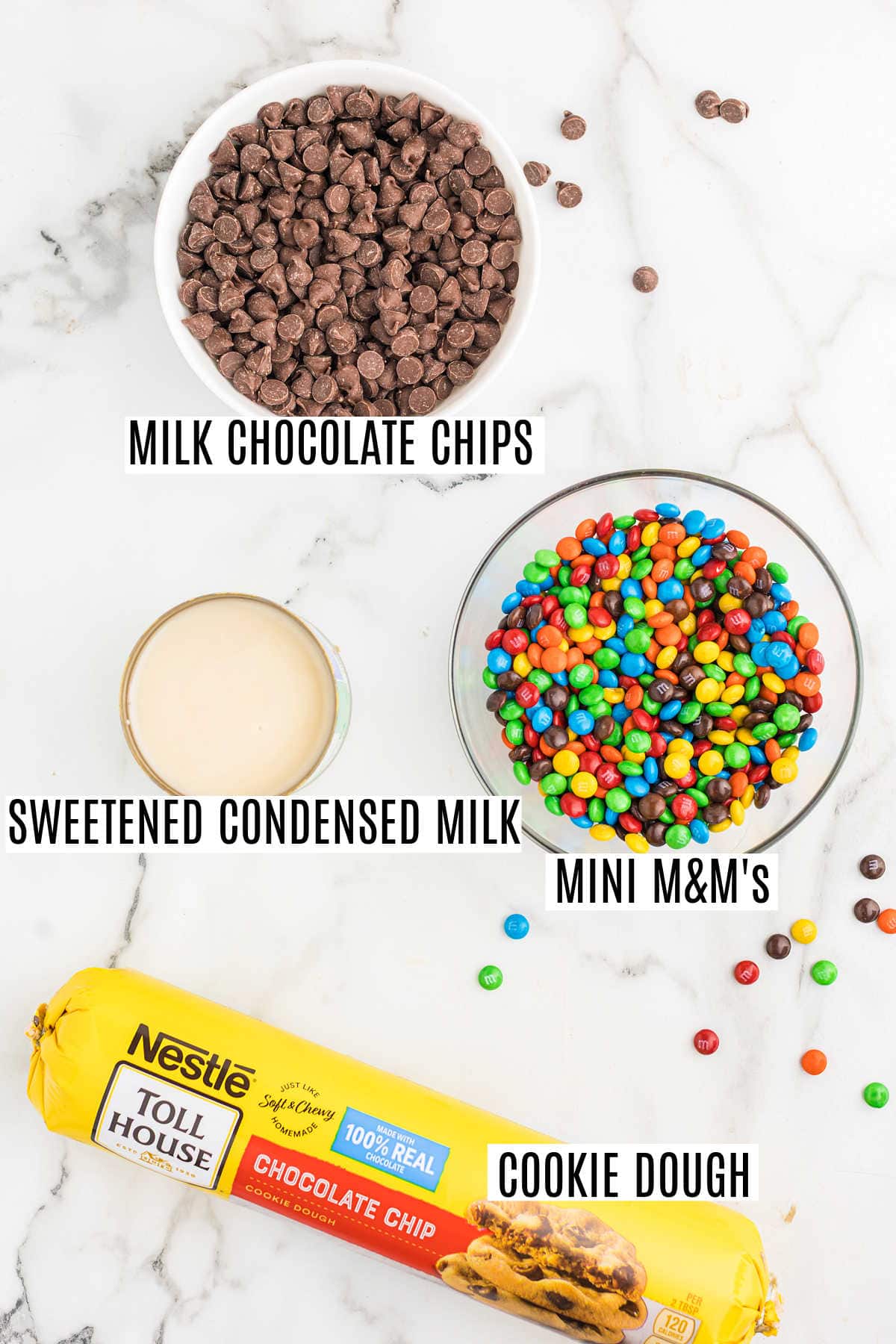 Bowls of milk chocolate chips, sweetened condensed milk, mini M&M’s, and a roll of chocolate chip cookie dough—all you need to whip up delicious rainbow fudge cookie bars.