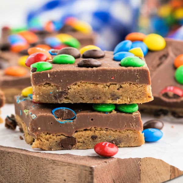 Two stacked rainbow fudge cookie bars topped with colorful candy-coated chocolates sit on a wooden board.
