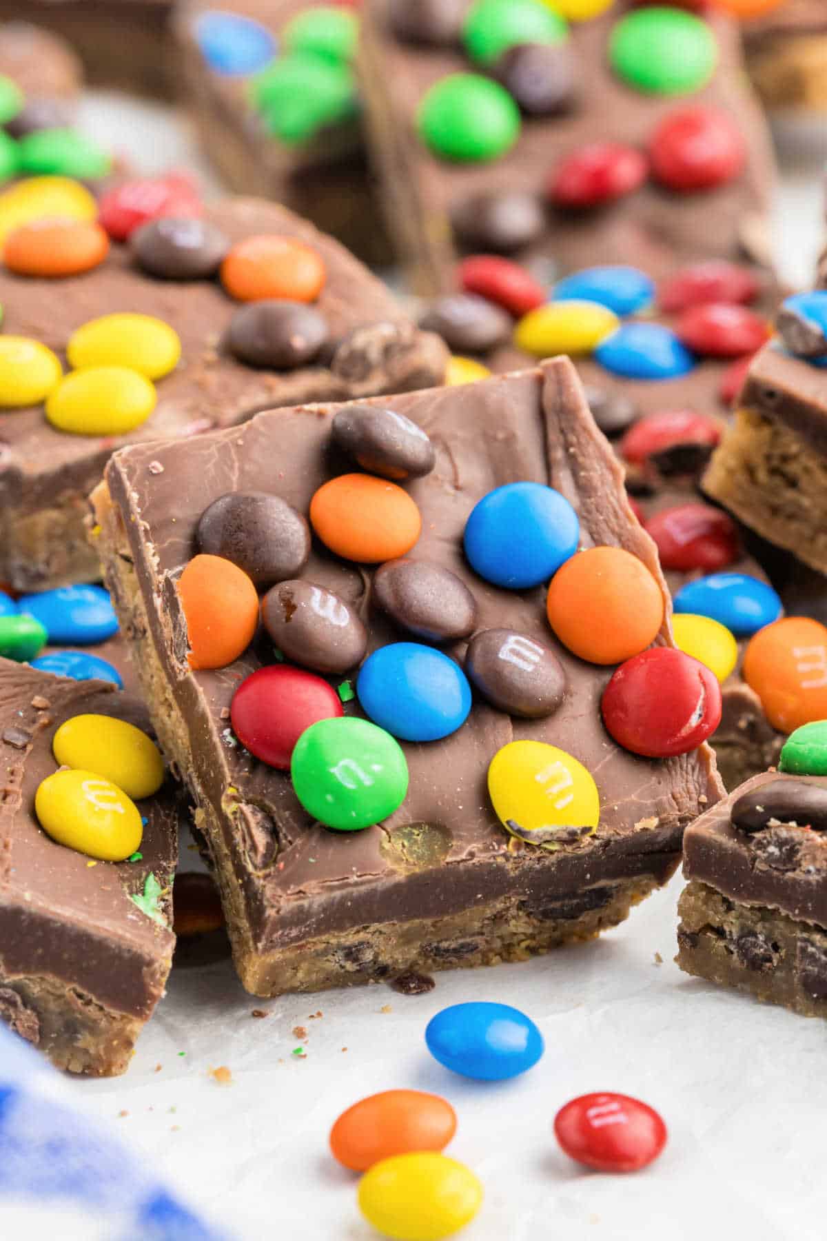 Close-up of rainbow fudge cookie bars, chocolate squares topped with colorful candy-coated chocolates, stacked on a white surface.