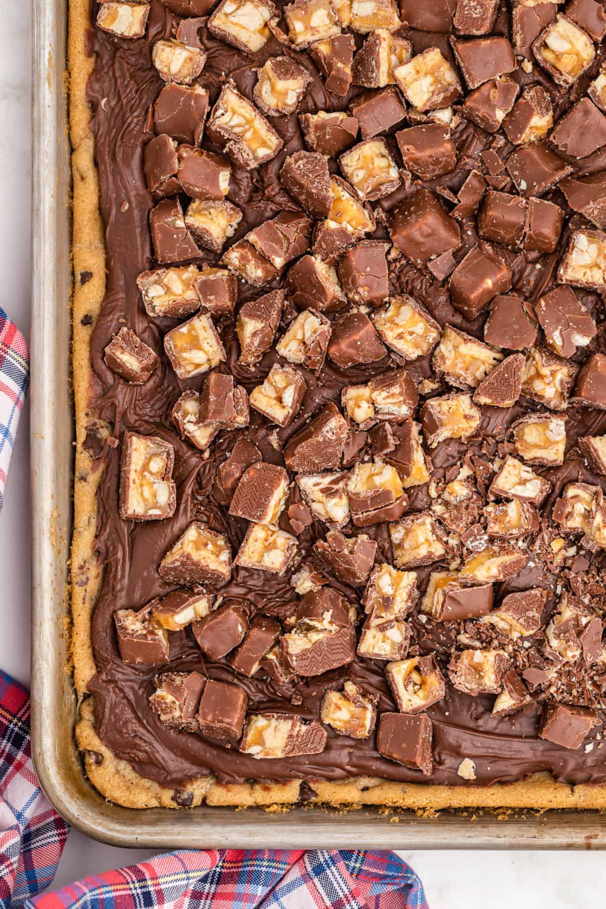 Baking sheet with chocolate chip cookie bars topped with snickers and fudge.