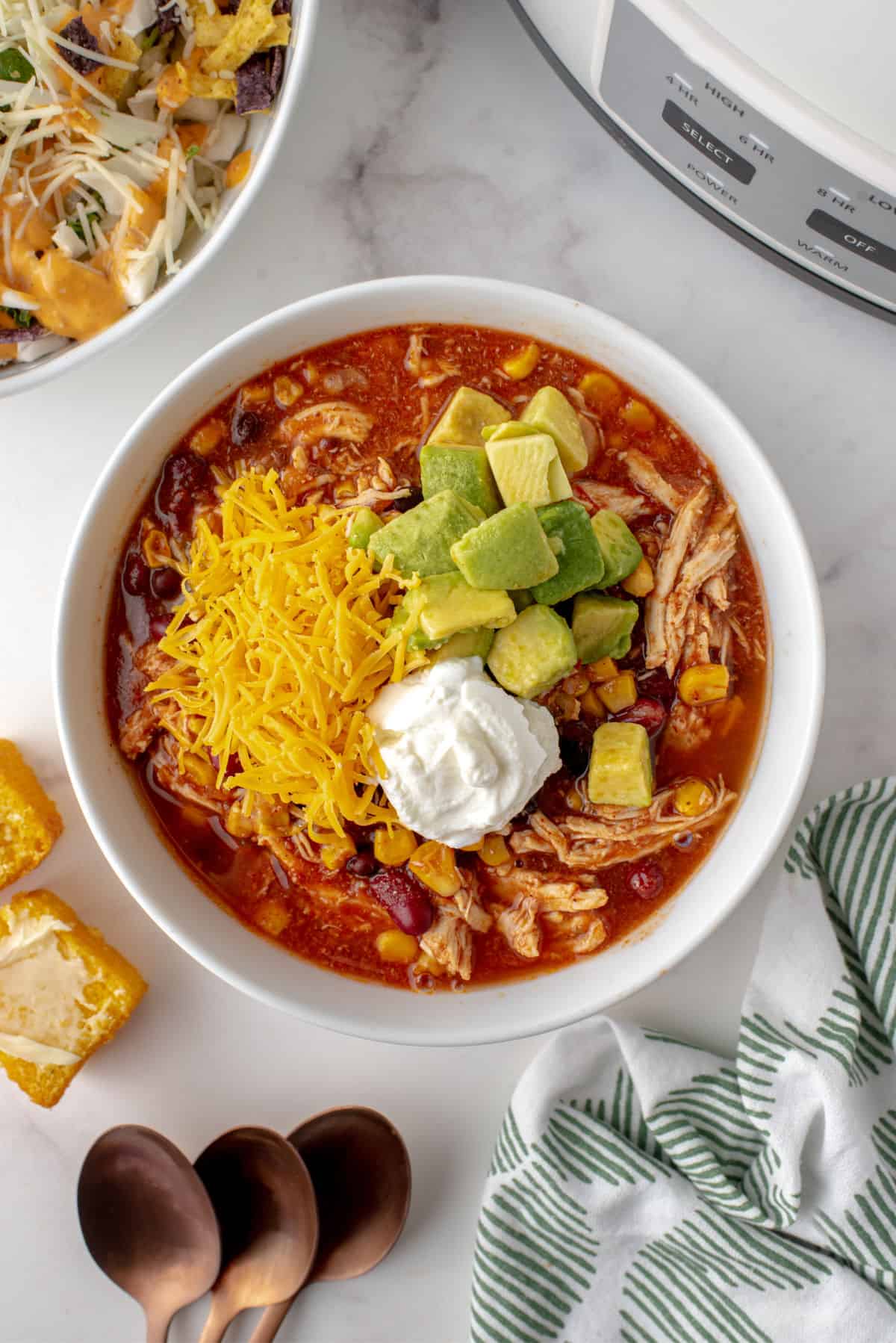 Chicken taco chili in a white bowl garnished with cheese, avocado, and sour cream.