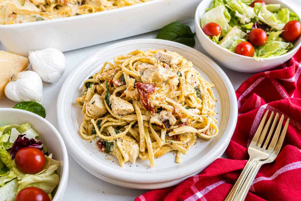 Plate of creamy pasta with chicken and sun-dried tomatoes, inspired by an Italian chicken casserole, served with salad and a red napkin on the side.