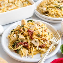 Plate of creamy pasta with chicken, sun-dried tomatoes, and spinach—an Italian chicken casserole twist—served with a gold fork on a white table.