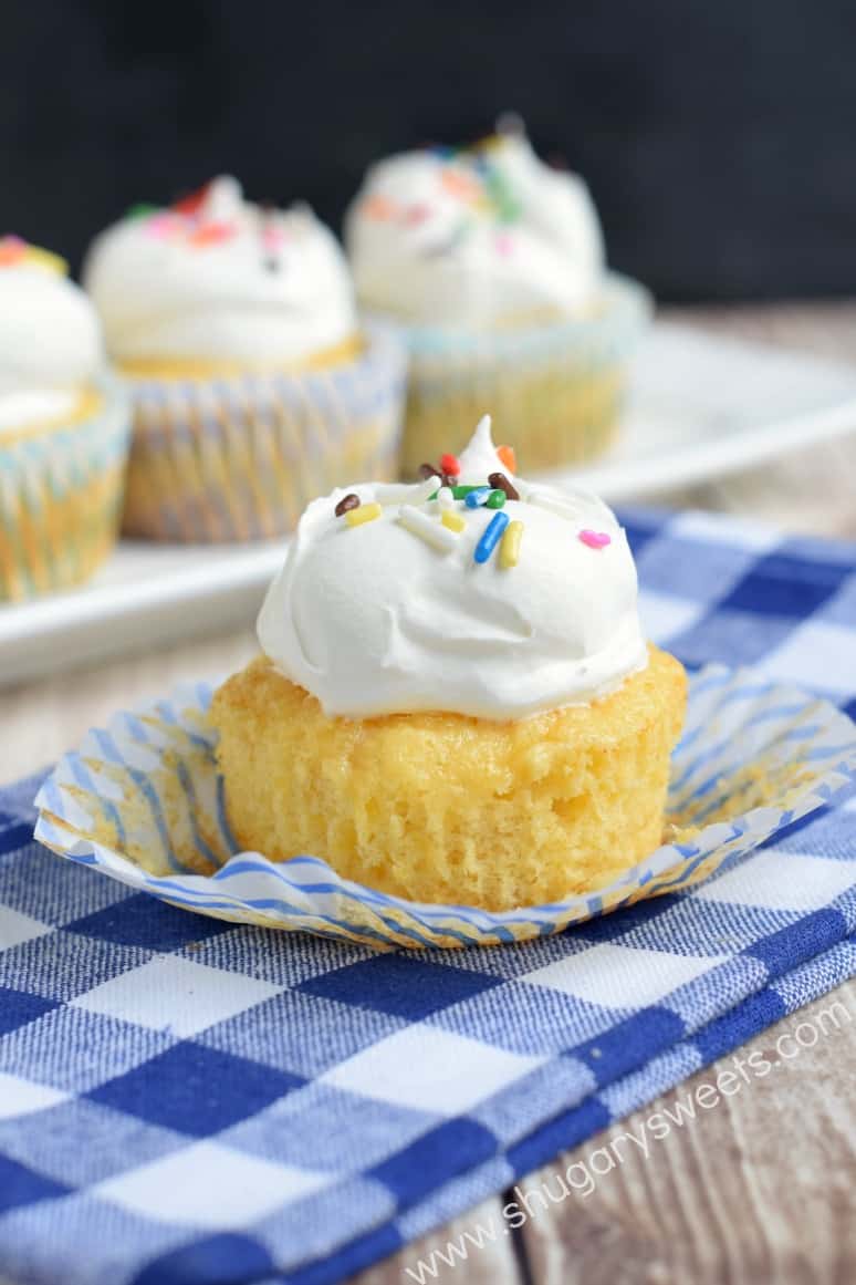 Pineapple cupcake topped with whipped cream and sprinkles unwrapped on a blue checkered napkin.