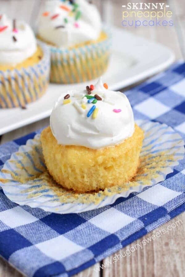 Pineapple cupcake on a blue checkered linen with the wrapper undone and topped with cool whip and sprinkles.