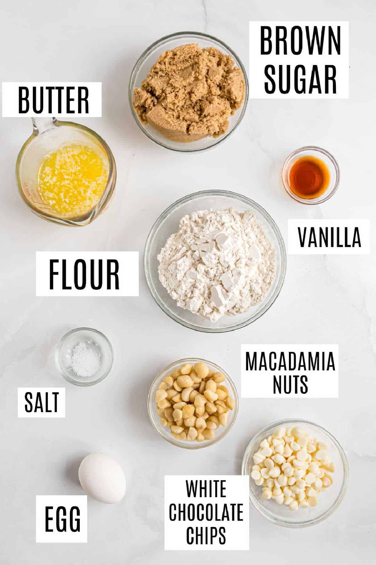 Bowls of brown sugar, flour, melted butter, vanilla, salt, egg, macadamia nuts, and white chocolate chips on a white surface—perfect ingredients for delicious macadamia nut blondies.