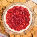 A wooden board boasts a glass dish brimming with cherry cheesecake dip, flanked by an assortment of cookies, pretzels, and graham crackers.