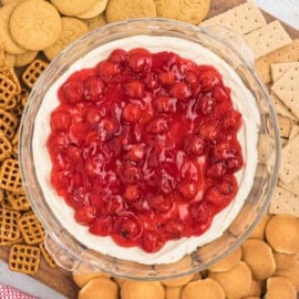 A wooden board boasts a glass dish brimming with cherry cheesecake dip, flanked by an assortment of cookies, pretzels, and graham crackers.