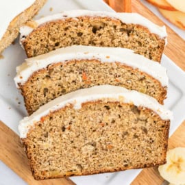 Slices of frosted banana-carrot bread on a white plate, with visible banana pieces and a wooden surface beneath.