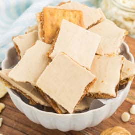 A bowl filled with square, frosted cracker snacks, reminiscent of peanut butter cracker toffee, sits on a wooden table.