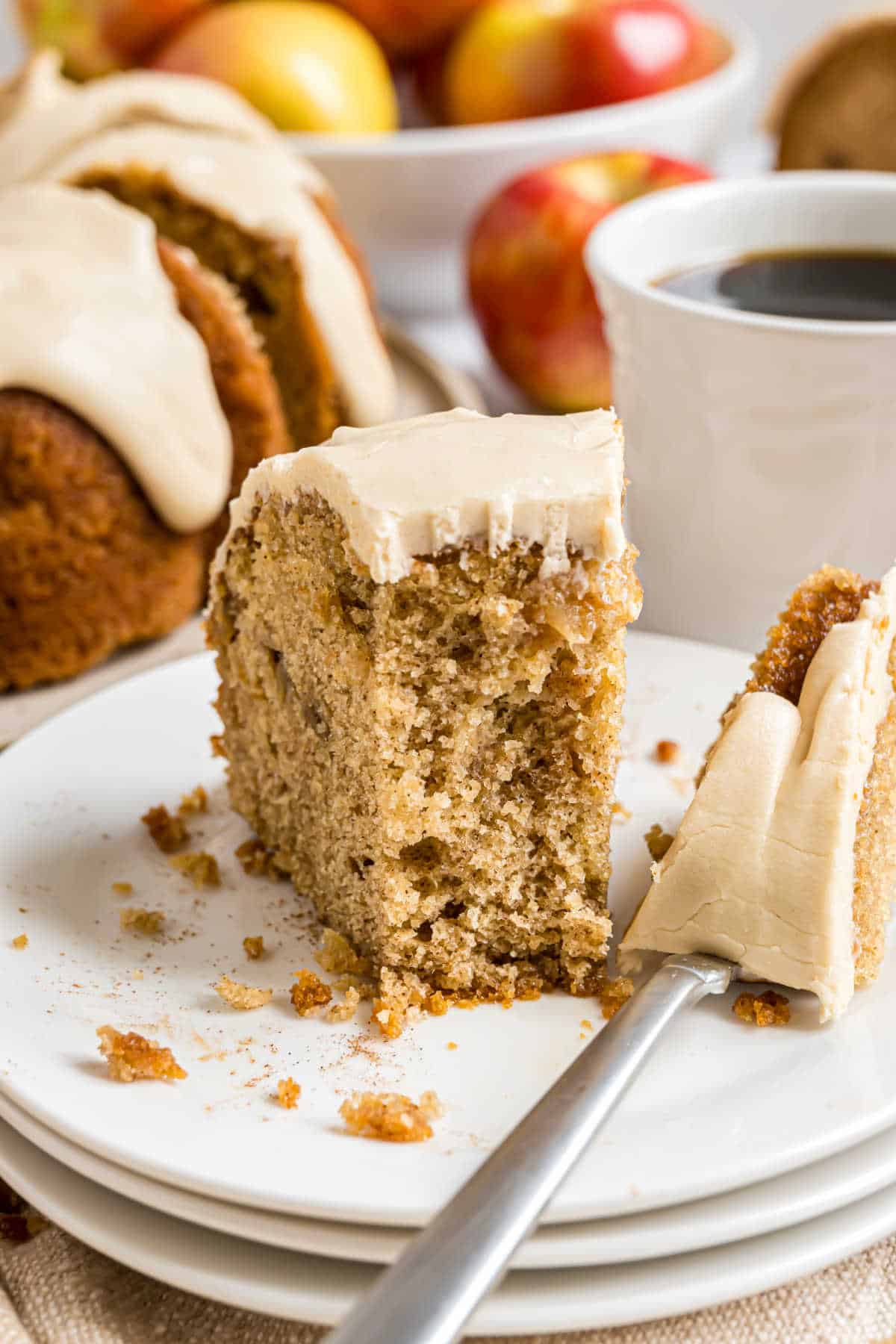 A slice of caramel apple bundt cake sits on a plate with a fork, while coffee and fresh apples add warmth to the background.