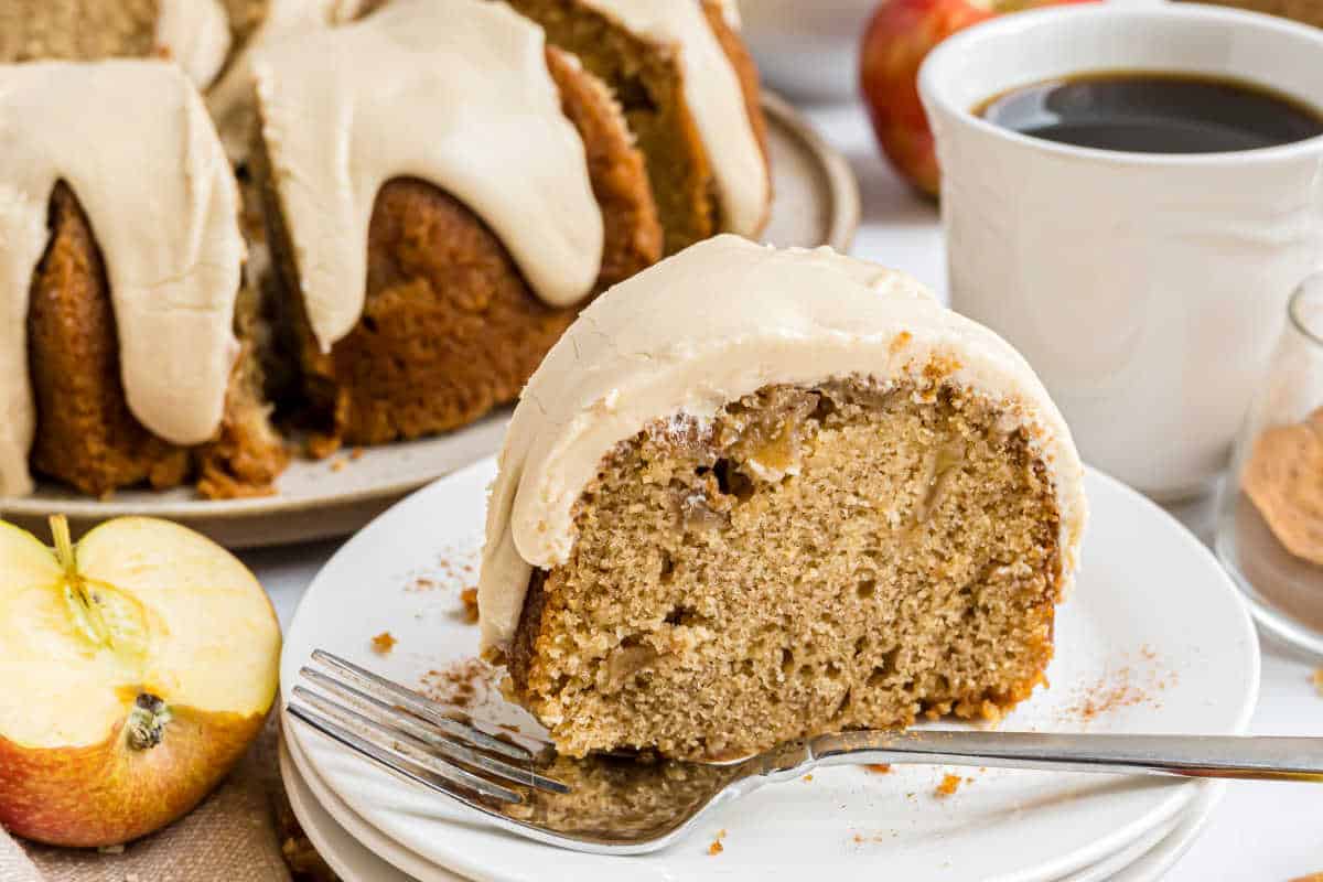 A slice of caramel apple bundt cake on a plate with a fork, coffee, and apples in the background.