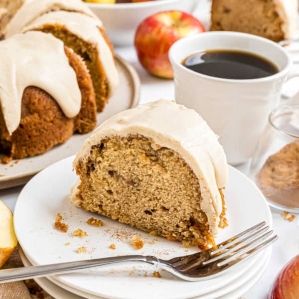 A slice of caramel apple bundt cake sits on a plate with a fork, coffee, and apples in the background.
