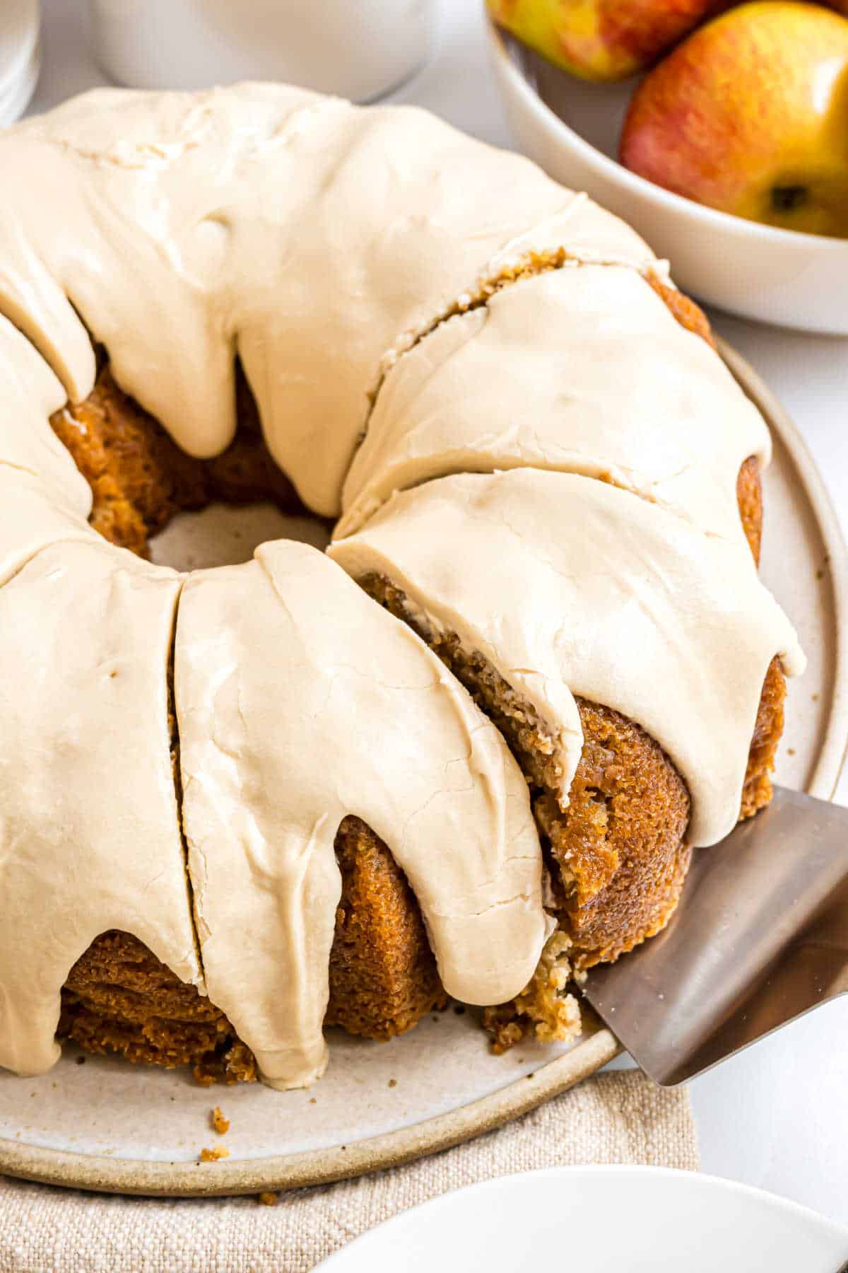 A sliced caramel apple bundt cake with beige icing sits on a plate, with fresh apples in a bowl in the background.