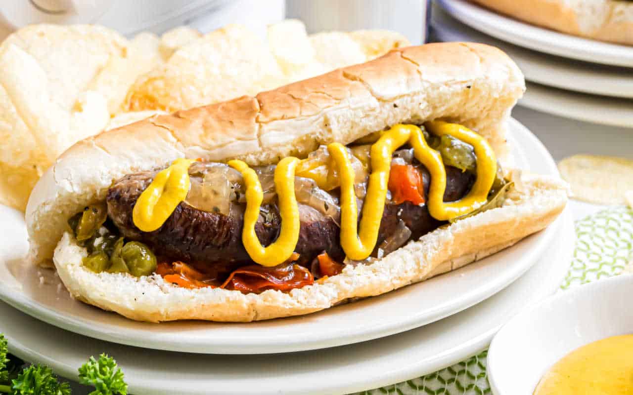 A slow cooker beer brat in a bun with mustard, peppers, and onions is served on a white plate alongside crispy potato chips.