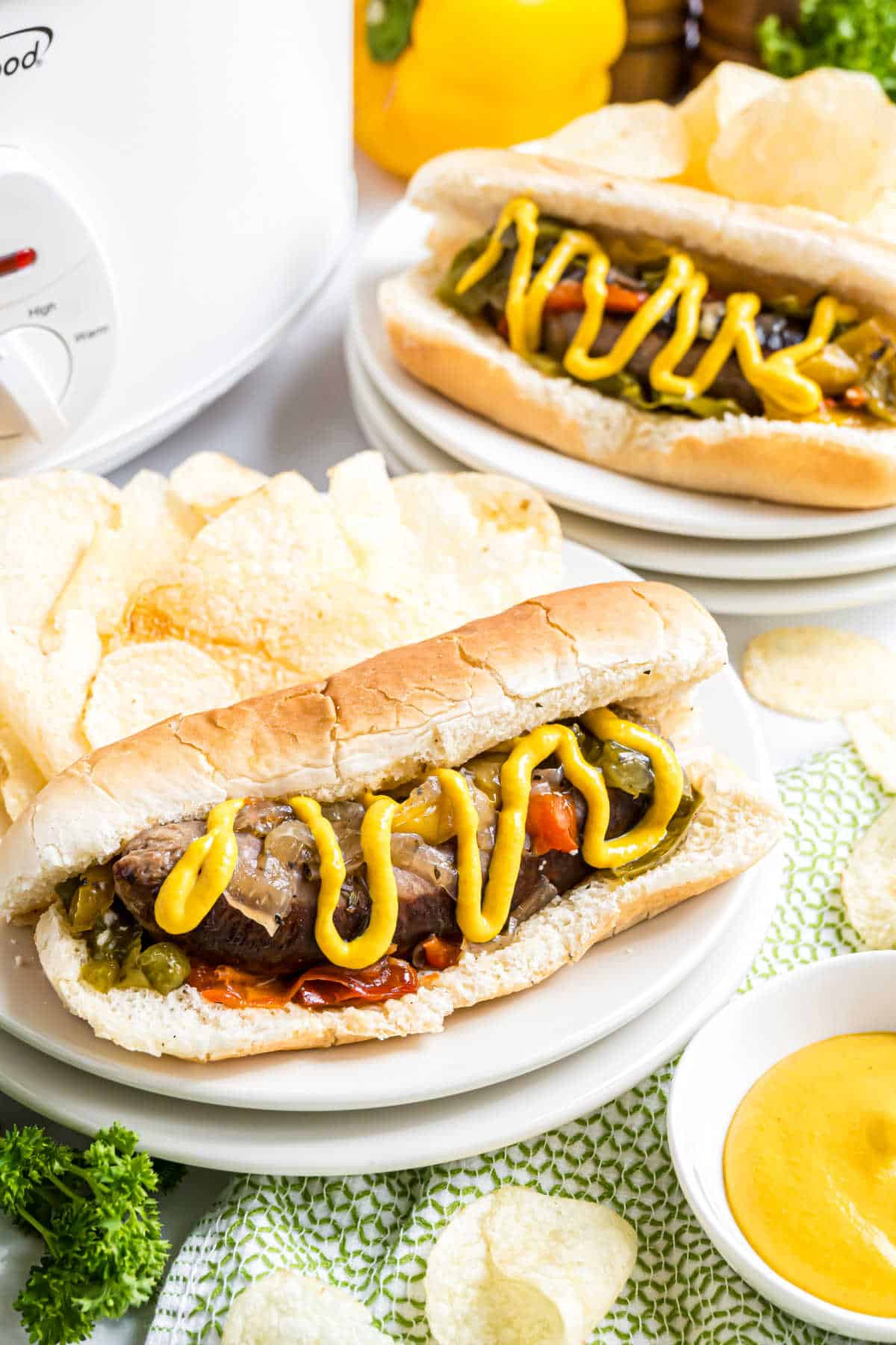 A hoagie roll with tender slow cooker beer brats, beef, peppers, and mustard, served with potato chips on a white plate.