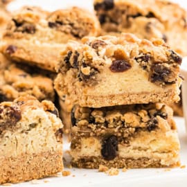 Close-up of crumbly oatmeal raisin cookie cheesecake bars with raisins, stacked on a white surface.