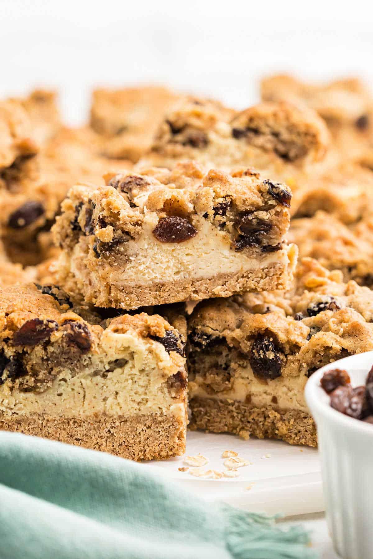 A close-up of stacked oatmeal raisin cookie cheesecake bars with a crumbly topping on a white surface.