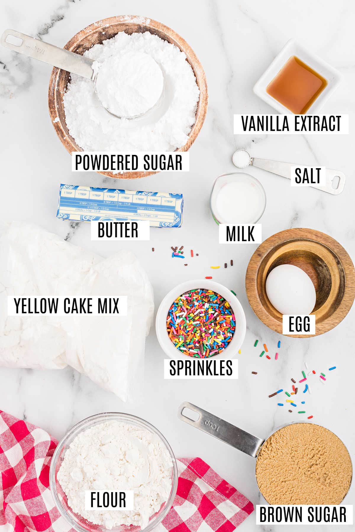 Baking ingredients on a counter labeled: powdered sugar, vanilla, salt, butter, milk, cake mix, sprinkles, egg, flour, brown sugar—everything you need to make delicious cake batter blondies.