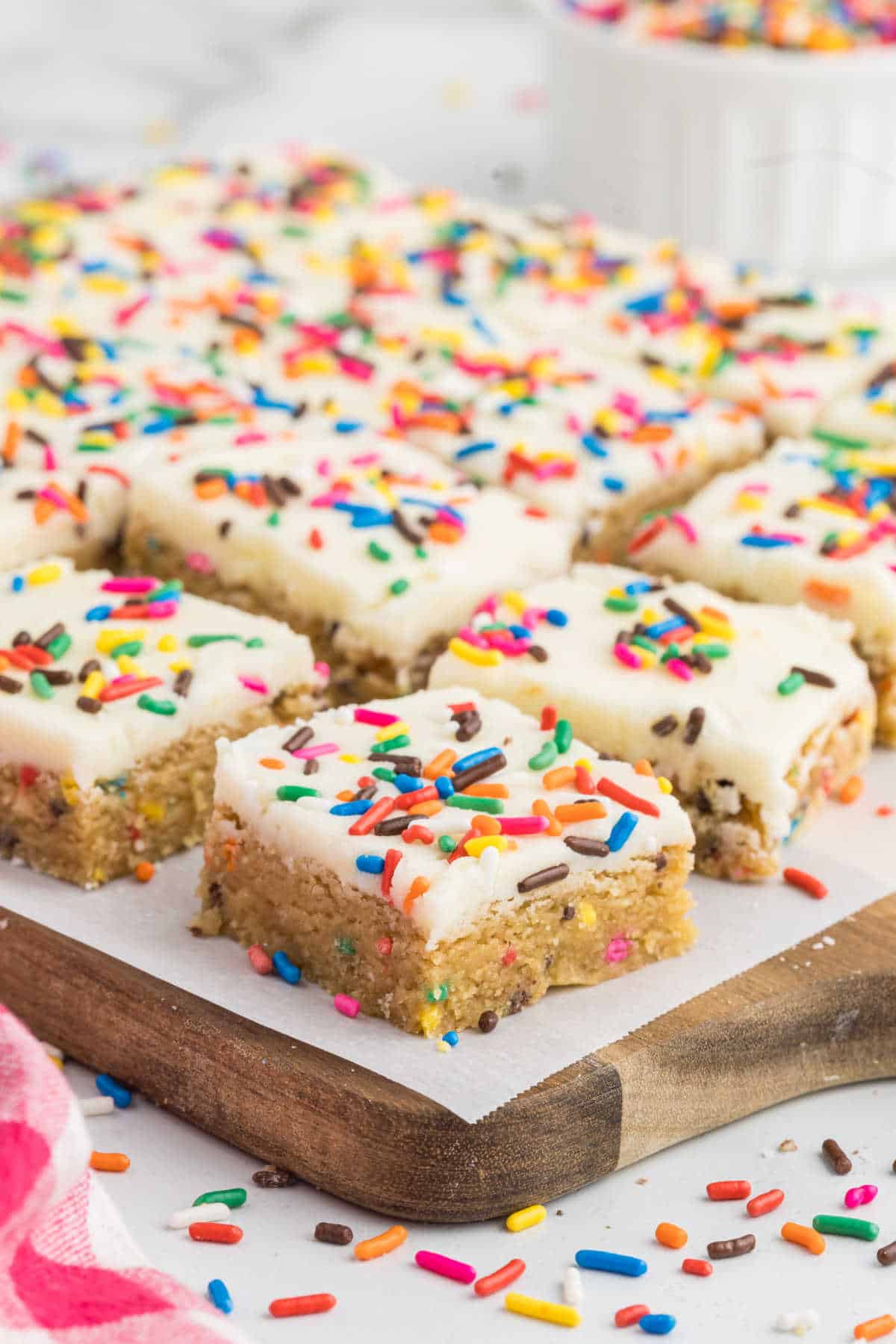 Frosted cake batter blondies with colorful sprinkles on a wooden board, cut into squares.
