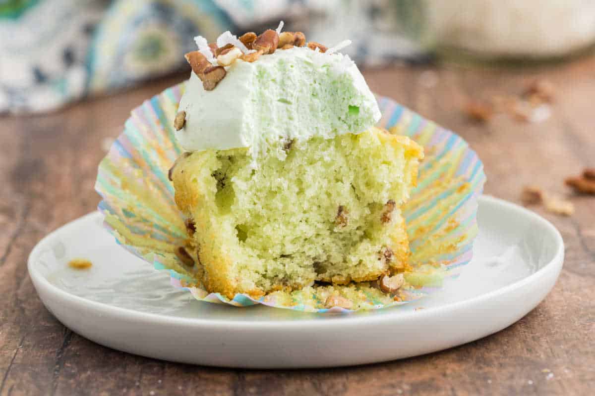 A half-eaten Watergate cupcake with green frosting and chopped nuts sits on a white plate.