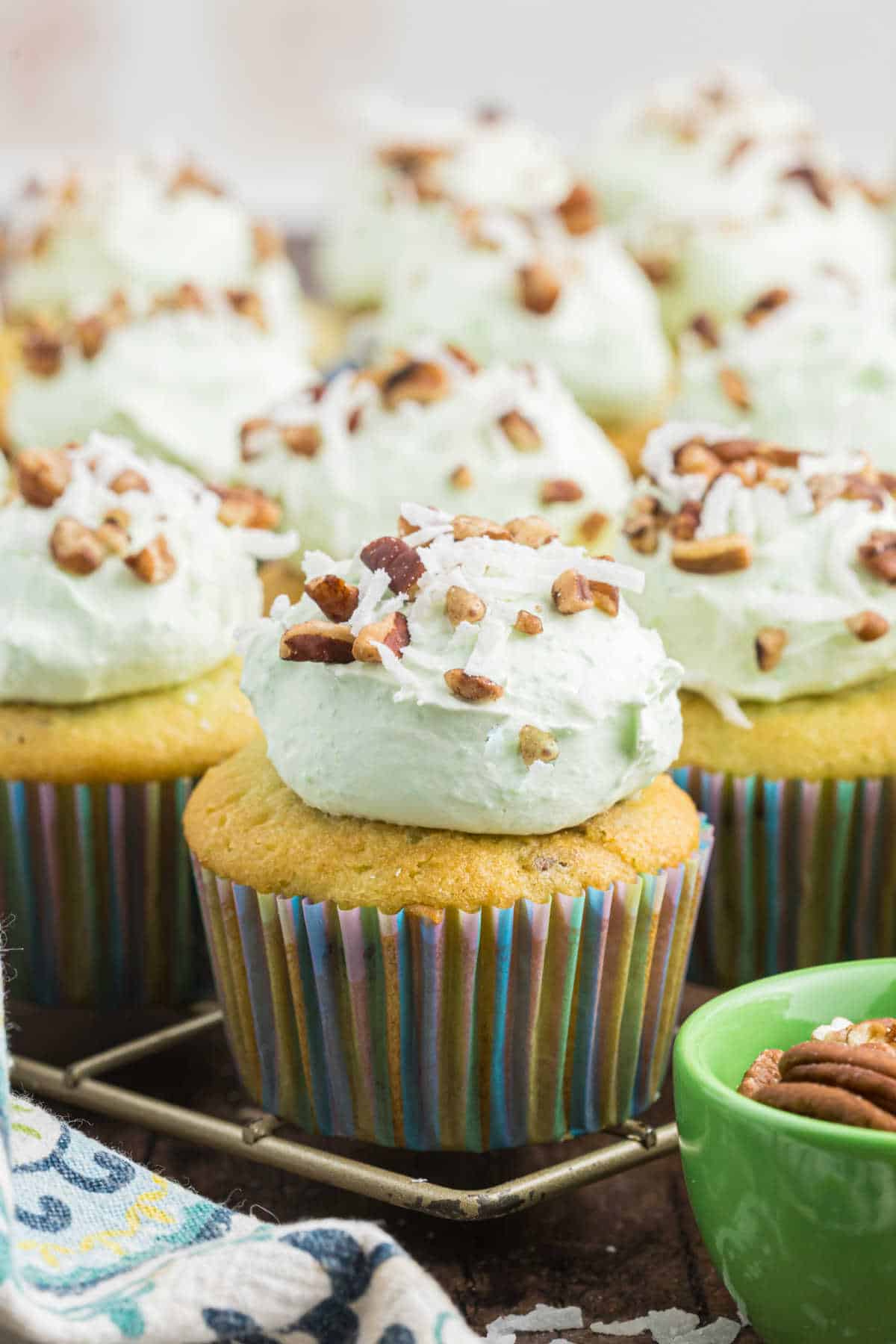 Watergate cupcakes with light green frosting are topped with chopped pecans and coconut, all in colorful wrappers.