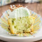 A Watergate cupcake with green frosting, chopped nuts, and a bite taken out, sits on a colorful wrapper.