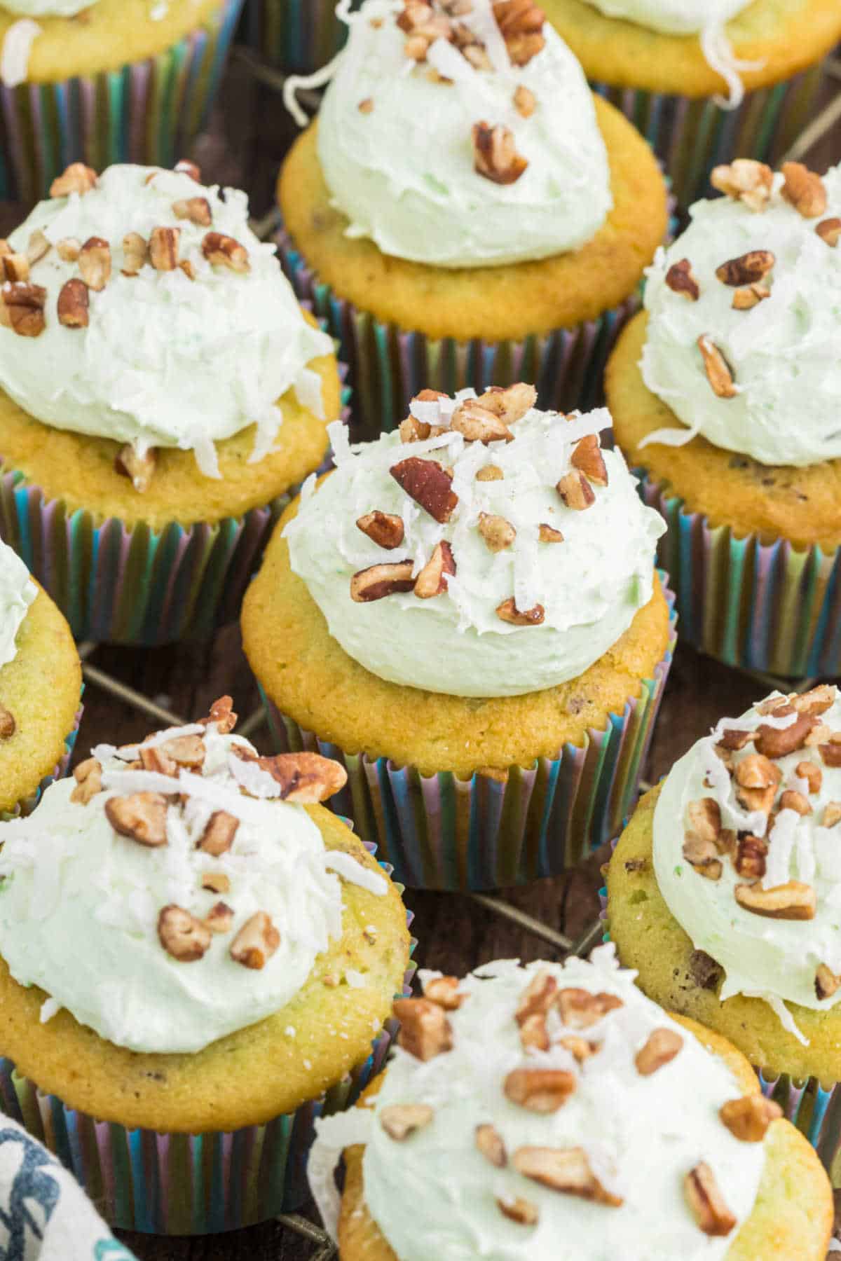 Watergate cupcakes with light green frosting, topped with chopped pecans and shredded coconut, in striped paper liners.