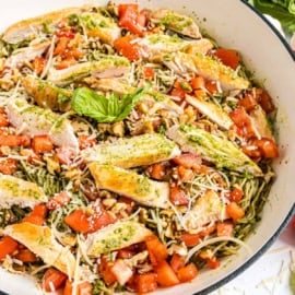 Spaghetti chicken pesto pasta topped with grilled chicken, diced tomatoes, pesto sauce, walnuts, and shredded parmesan in a white bowl.