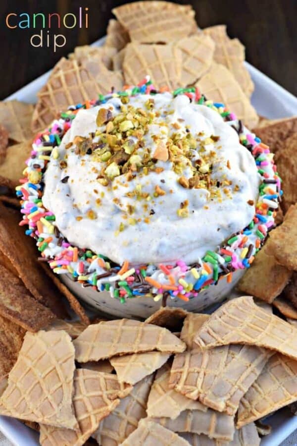 Cannoli dip in a bowl lined with sprinkles, topped with pistachios, and surrounded by waffle cone pieces.